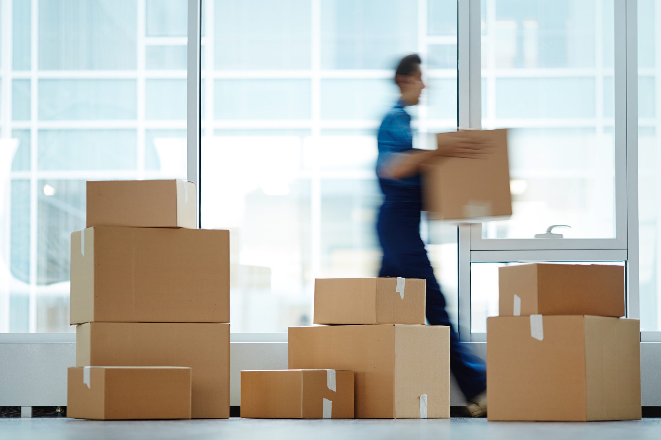 A worker carrying a cardboard box among stacked boxes in a warehouse.